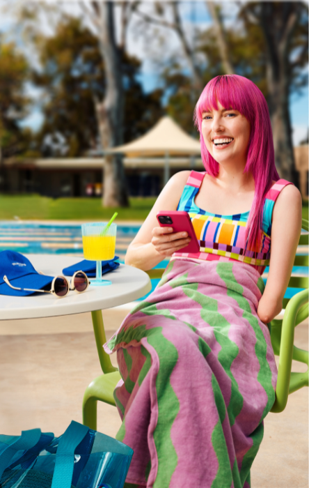 A smiling person with pink hair sits by a pool, wrapped in a colorful towel, using a phone. A drink and sunglasses are on the table